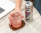 Person holding a Posca Hydrate Grape in a glass with a can on a table next to a laptop.