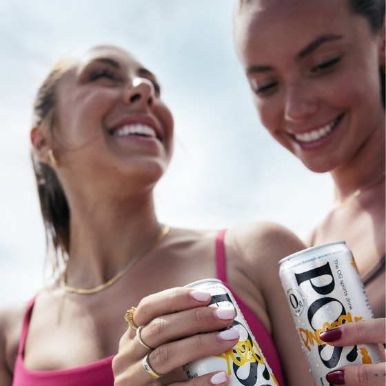 Two women holding cans of Posca Hydrate against a clear sky.