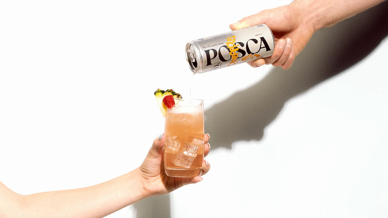 One hand holds a glass with ice and garnish whilst another hand pours Posca Hydrate Pineapple from a can, against a white background