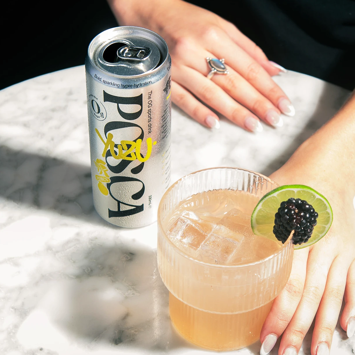 Person holding a glass of a Posca Hydrate Yuzu with a lime and blackberry garnish, next to a can on a marble surface.